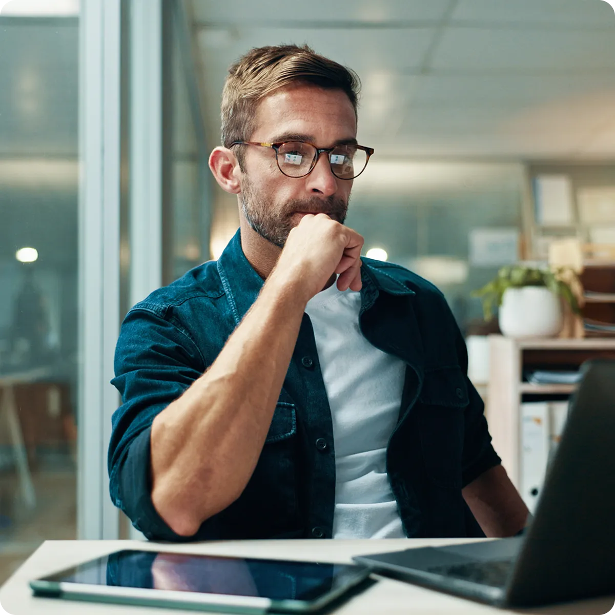 Man wearing glasses and a denim shirt, thoughtfully looking at a laptop in a modern office setting.