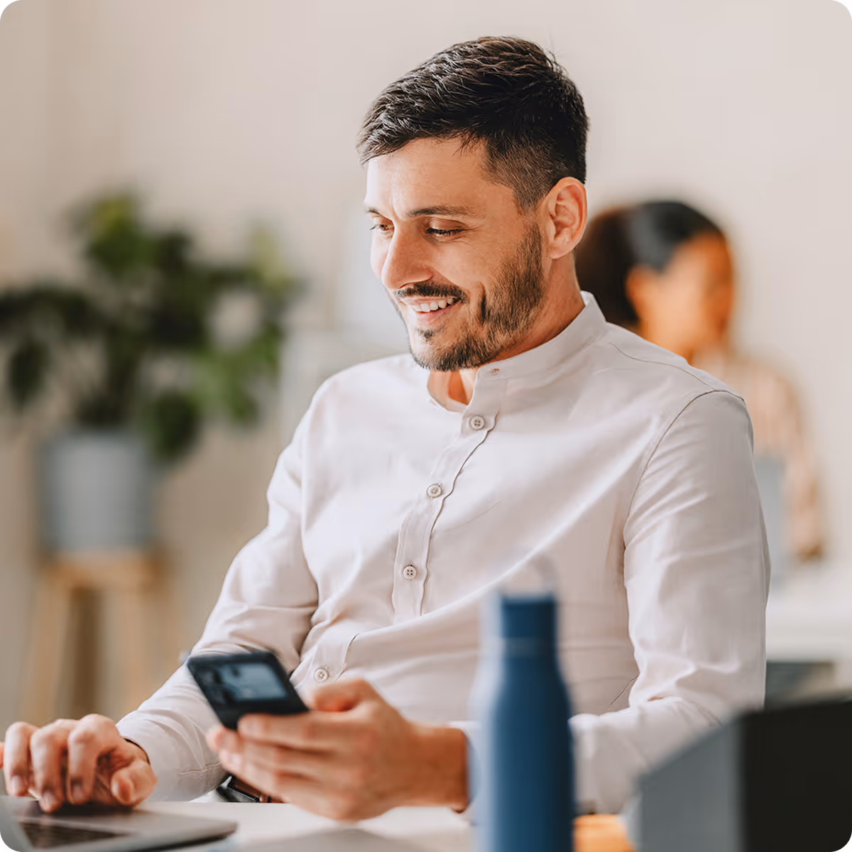 Smiling man in white shirt looking at his smartphone while sitting at a desk with a laptop and blue water bottle.