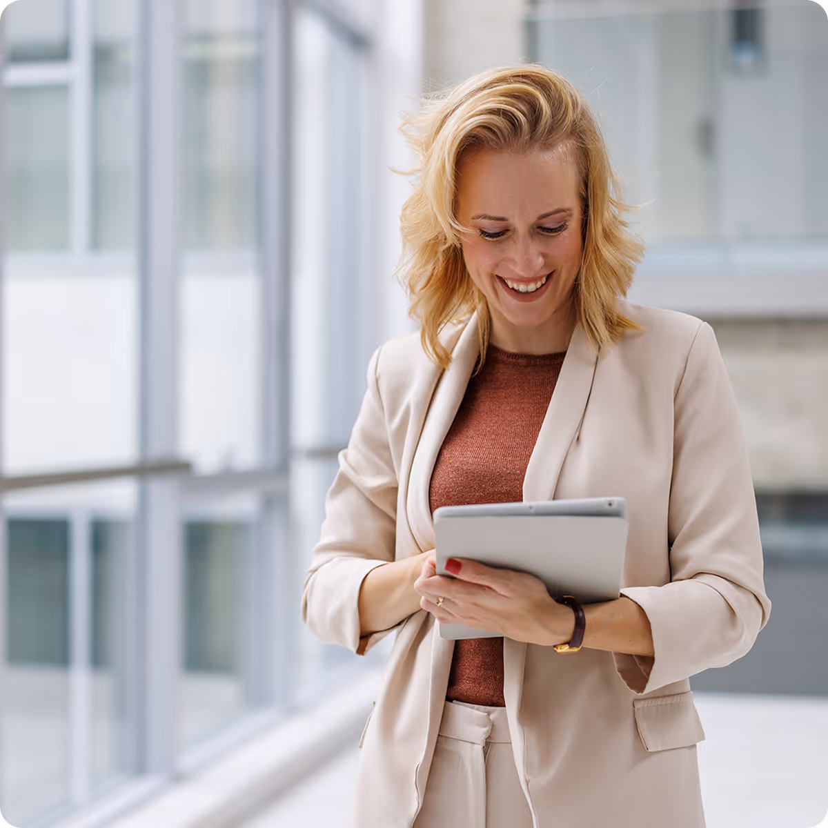 Smiling blonde woman in a beige suit holding and looking at a tablet in a modern office setting.