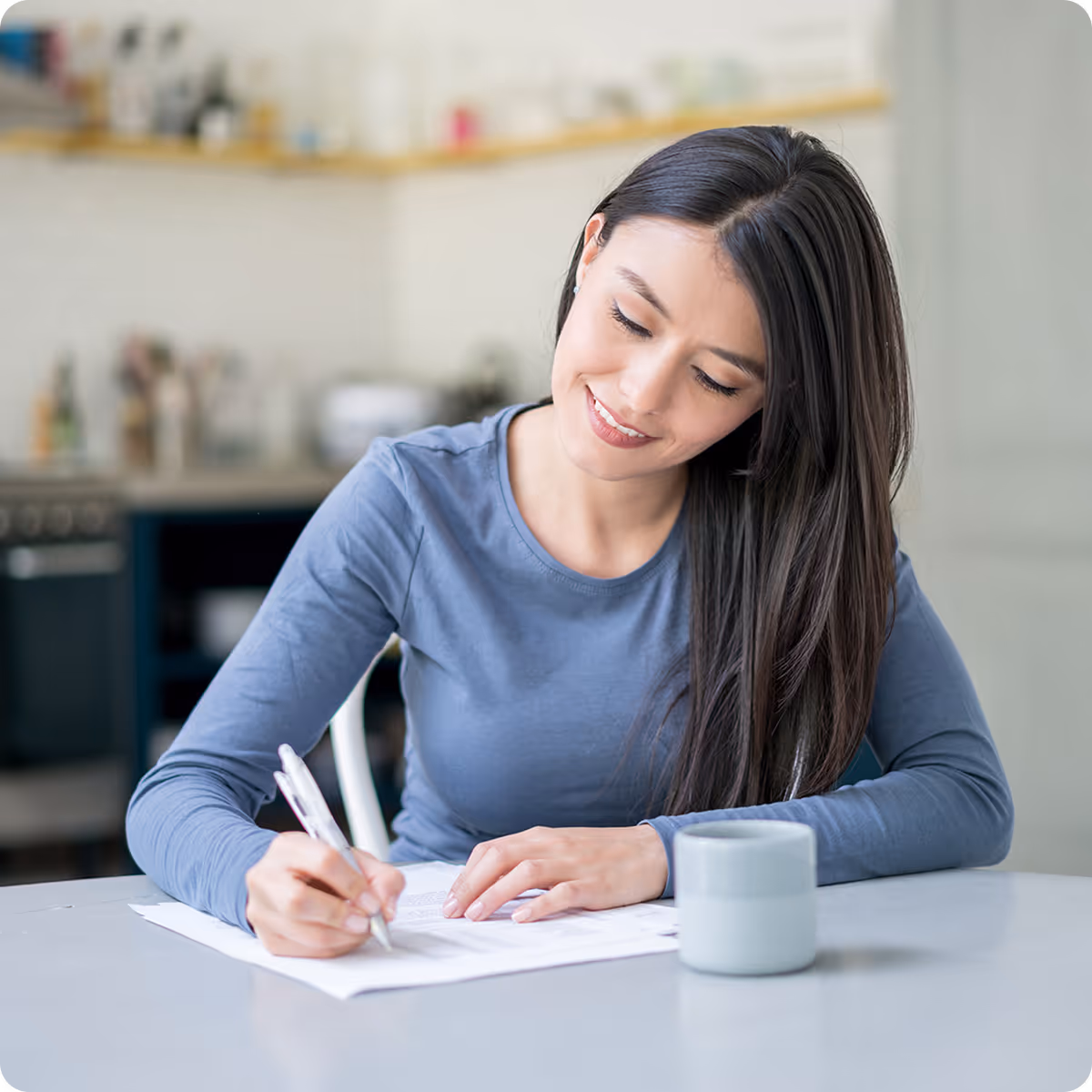 Smiling woman with long dark hair writing on paper at a table with a mug nearby in a kitchen.