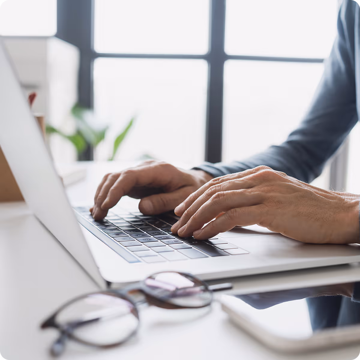 Person typing on a laptop keyboard at a bright workspace with glasses and smartphone nearby.
