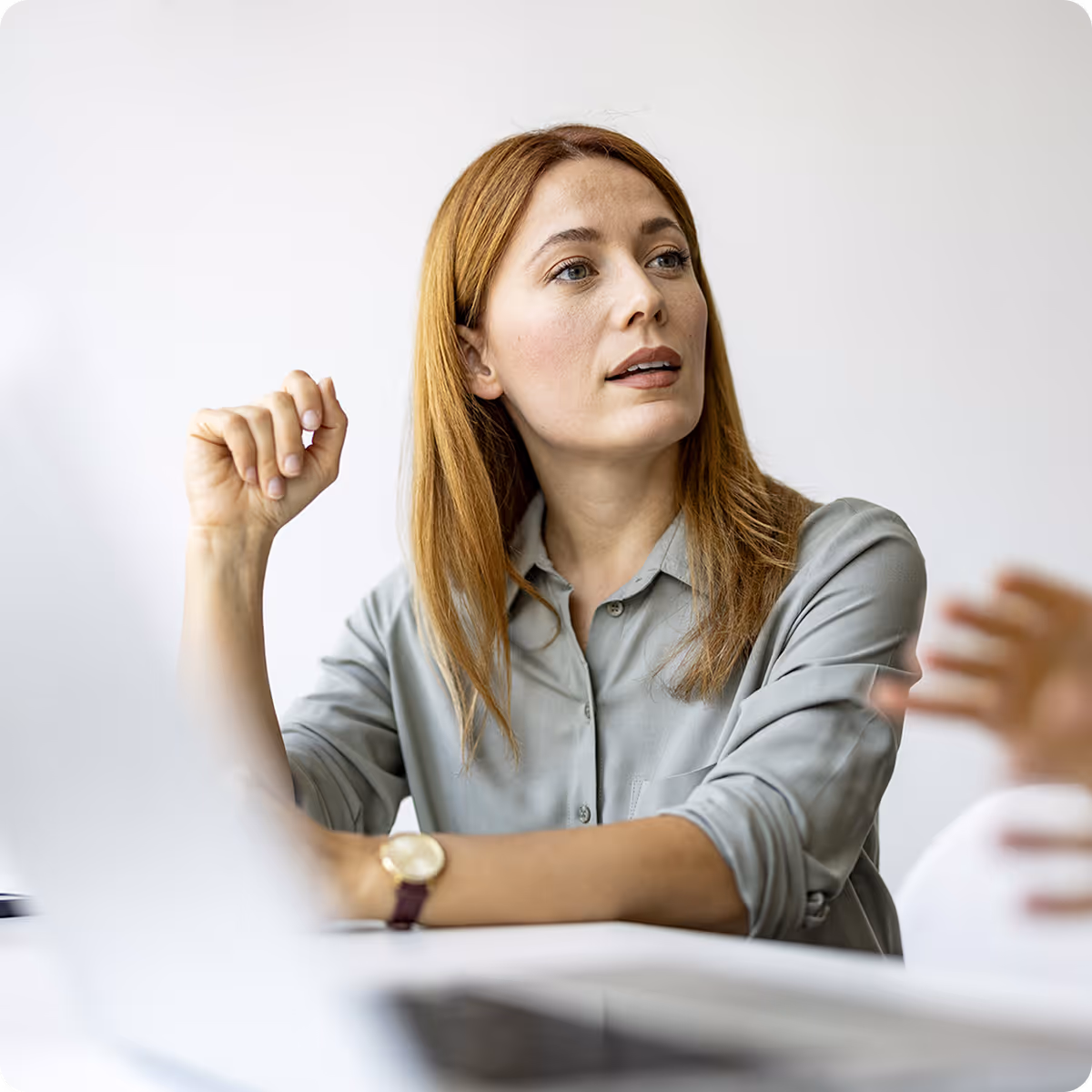 Woman with red hair wearing a gray shirt attentively listening and gesturing in a discussion.