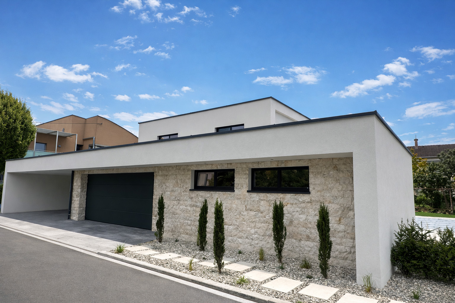 Modern single-story house with white walls, stone facade, black garage door, and tall narrow bushes along a gravel path.