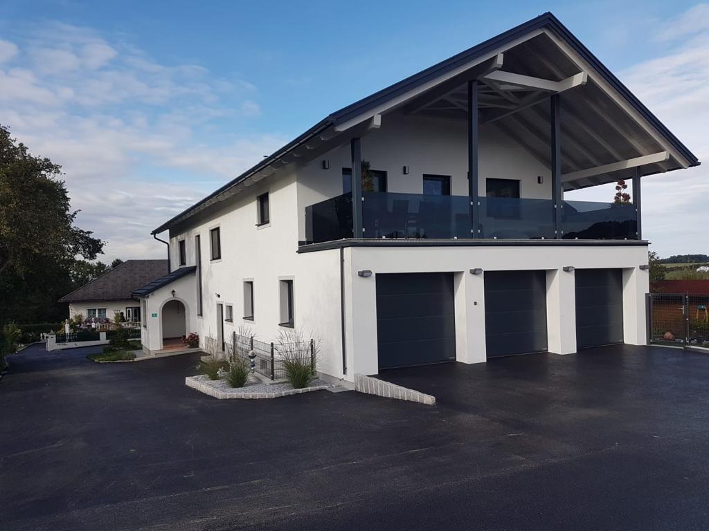 Modernes zweistöckiges weißes Haus mit drei schwarzen Garagentoren und einem überdachten Balkon mit Glasgeländer.