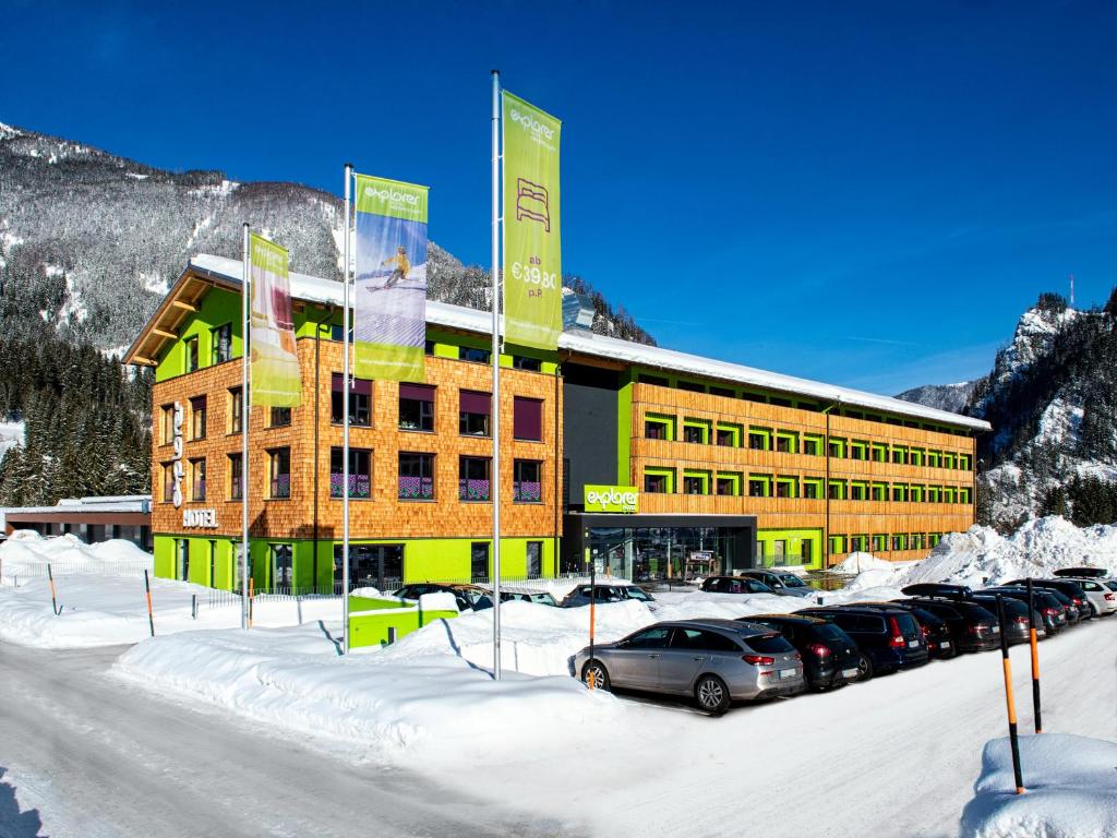 Explorer Hotel mit holz- und grünfarbener Fassade, eingeschneitem Parkplatz und Bergen im Hintergrund unter klarem blauem Himmel.