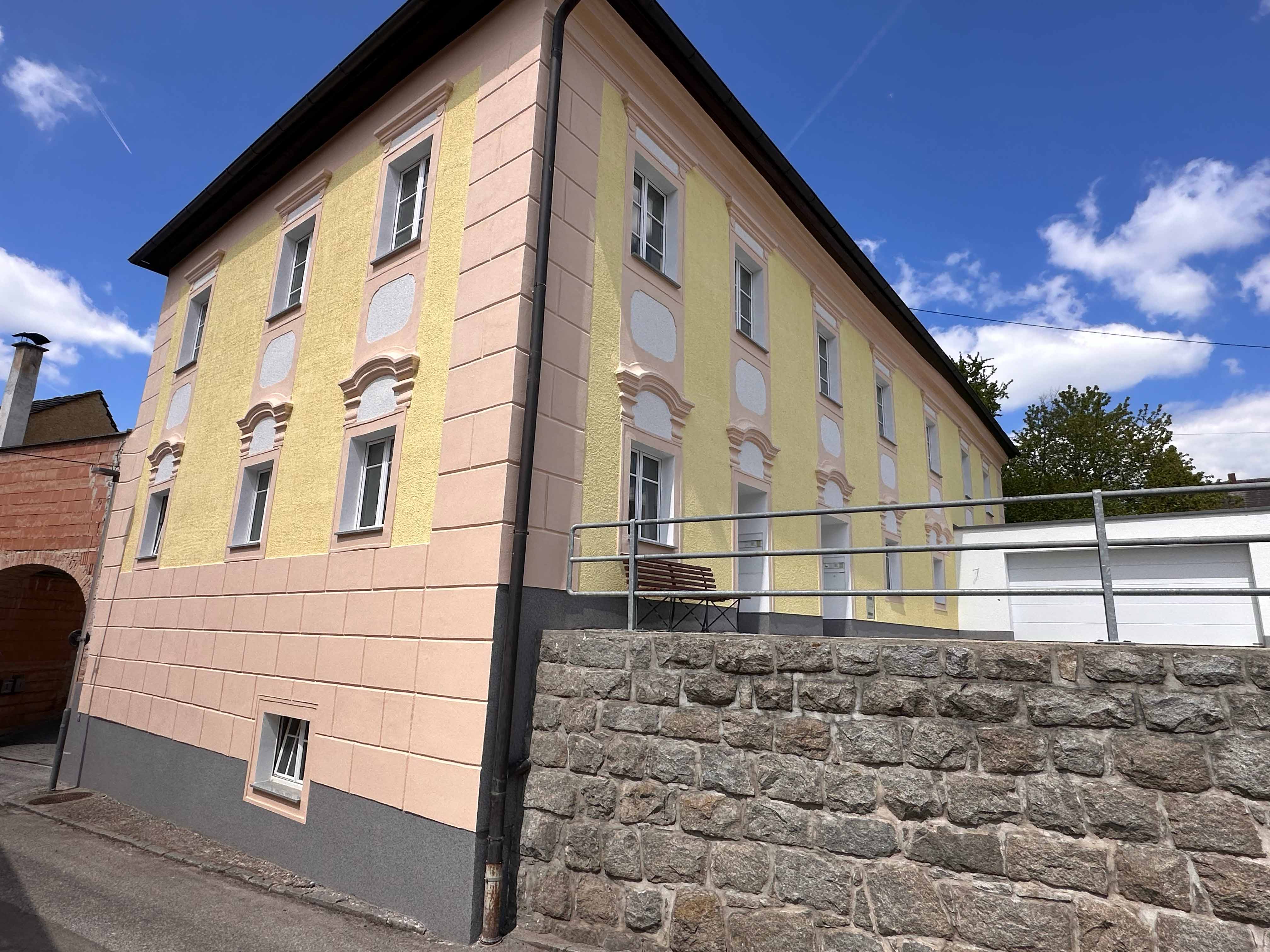 Mehrstöckiges Haus mit gelb-beiger Fassade, weißen Fenstern und Steinmauer im Vordergrund unter blauem Himmel.