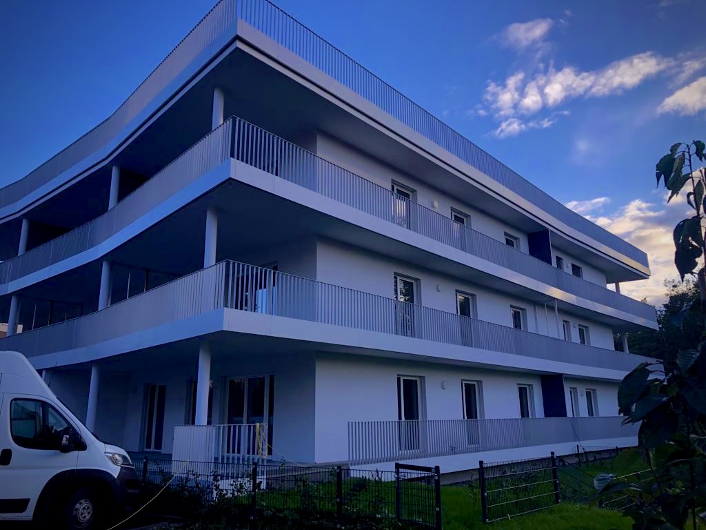 Modernes, dreistöckiges Wohngebäude mit weißen Balkonen und Geländern unter klarem blauem Himmel.