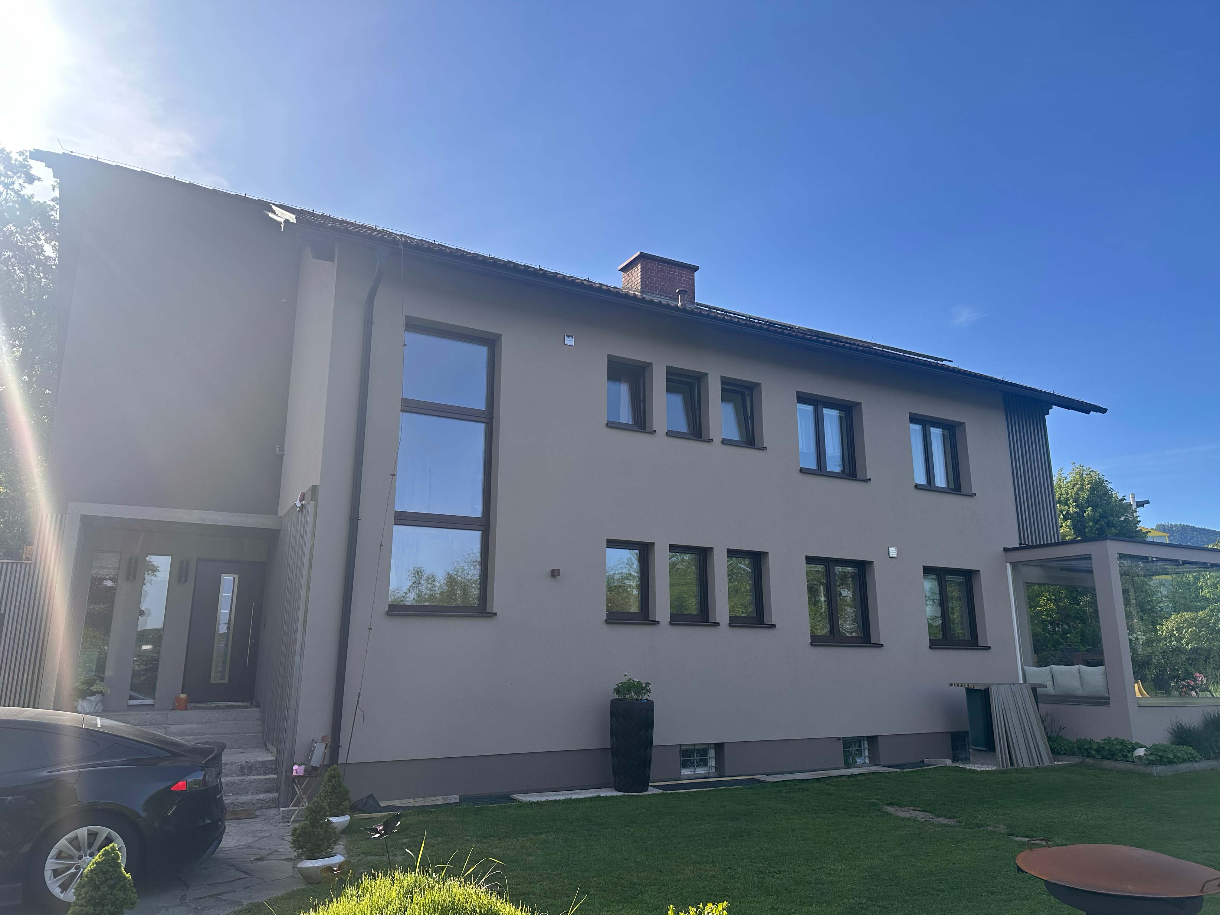 Modernes zweistöckiges Haus mit hellgrauer Fassade, mehreren Fenstern, einem kleinen Garten und einem schwarzen Auto im Einfahrtsbereich bei sonnigem blauem Himmel.