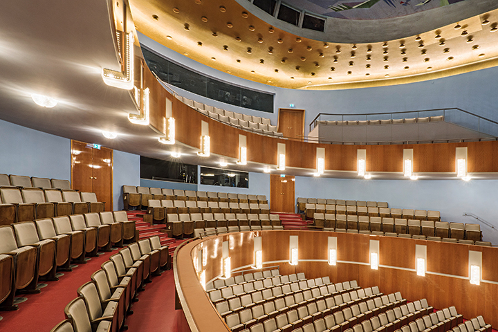 Innenansicht eines Theater- oder Konzertsaals mit beige gepolsterten Sitzen, roten Teppichböden und warmen Deckenbeleuchtungen.