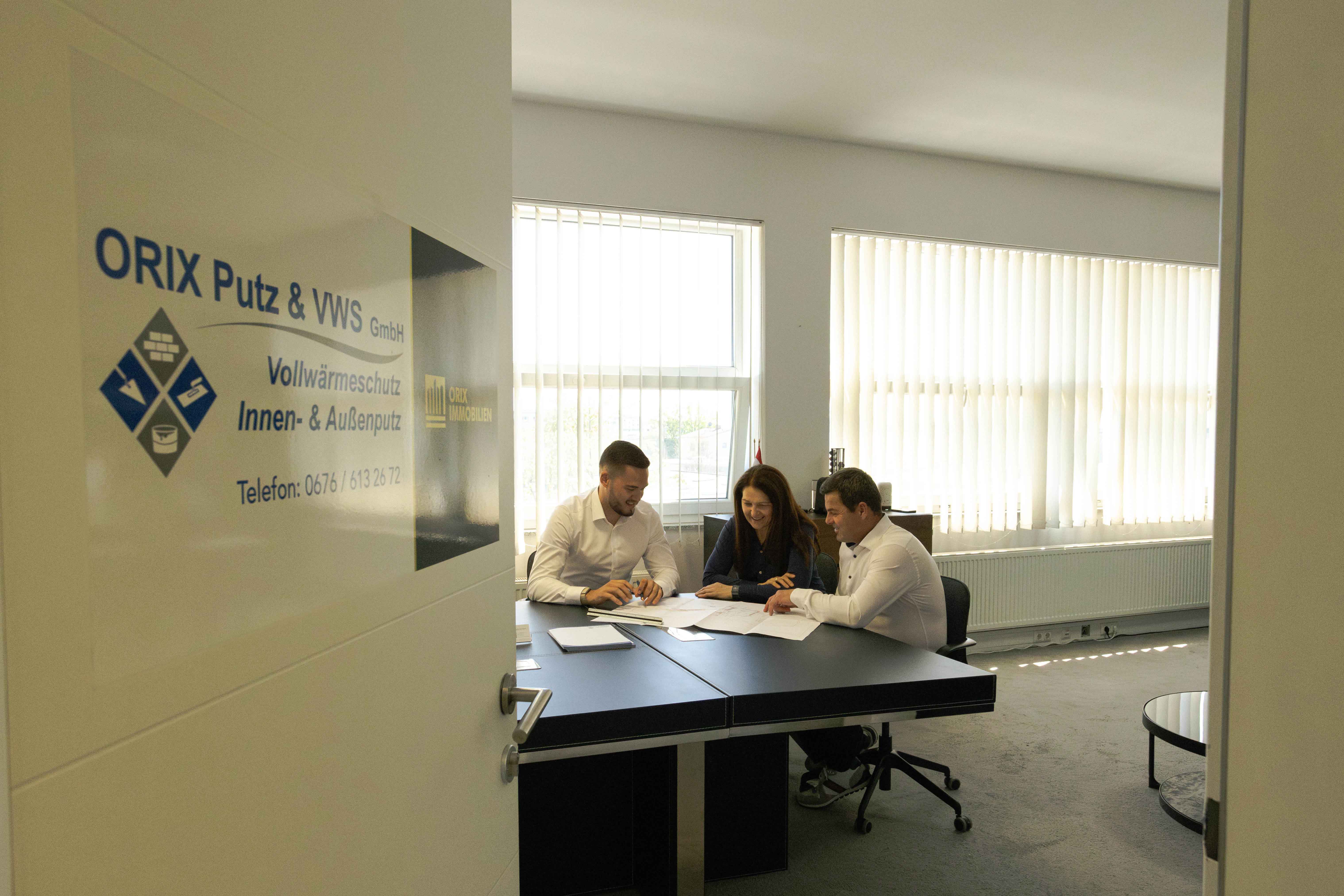 Drei Personen sitzen an einem Tisch in einem Büro und besprechen Unterlagen, neben der Tür ist ein Schild mit der Aufschrift ORIX Putz & VWS GmbH.