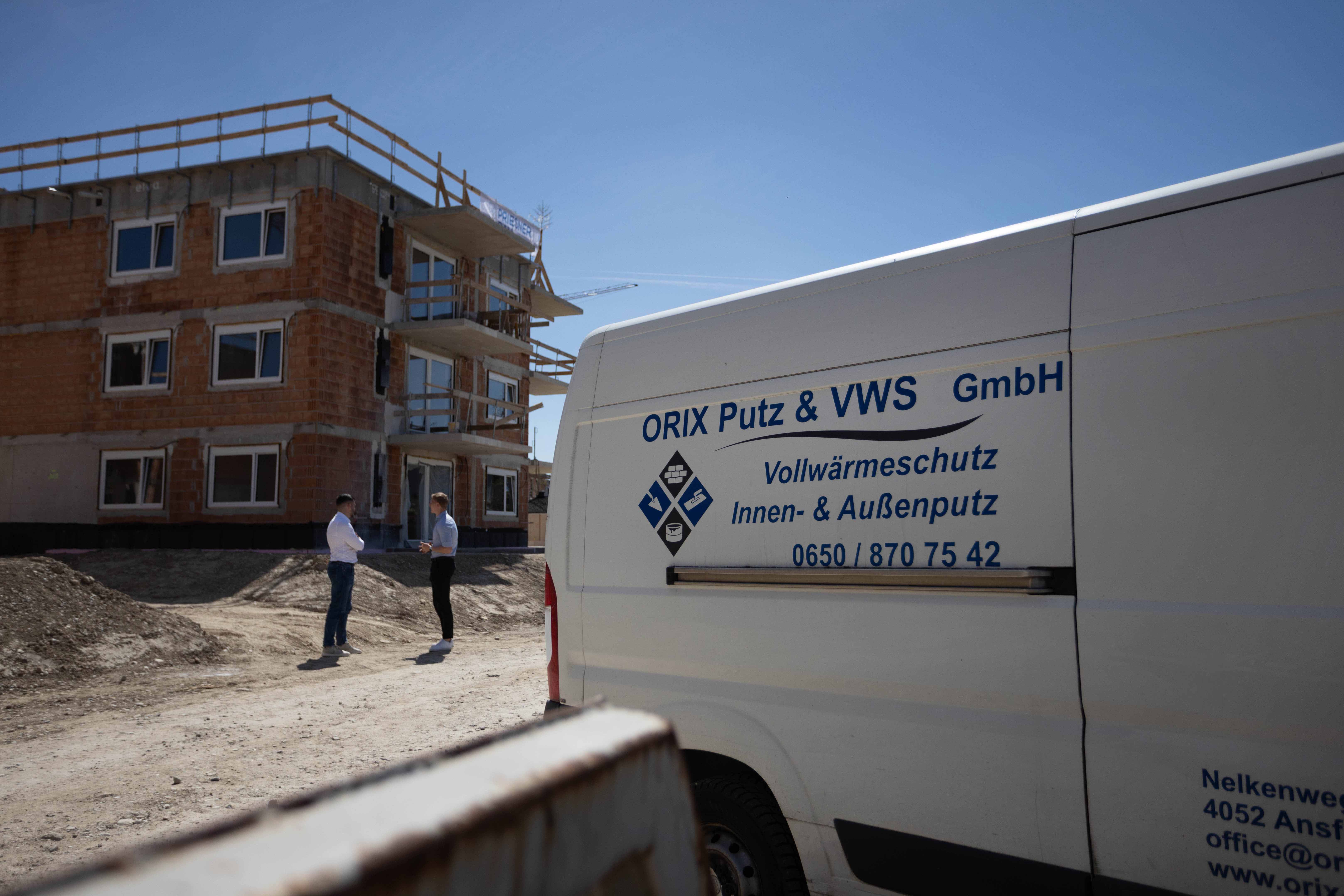 Bauarbeiter und Architekt im Gespräch vor einem Rohbau, weißer Firmenwagen von ORIX Putz & VWS GmbH mit Werbung für Vollwärmeschutz und Putzarbeiten im Vordergrund.