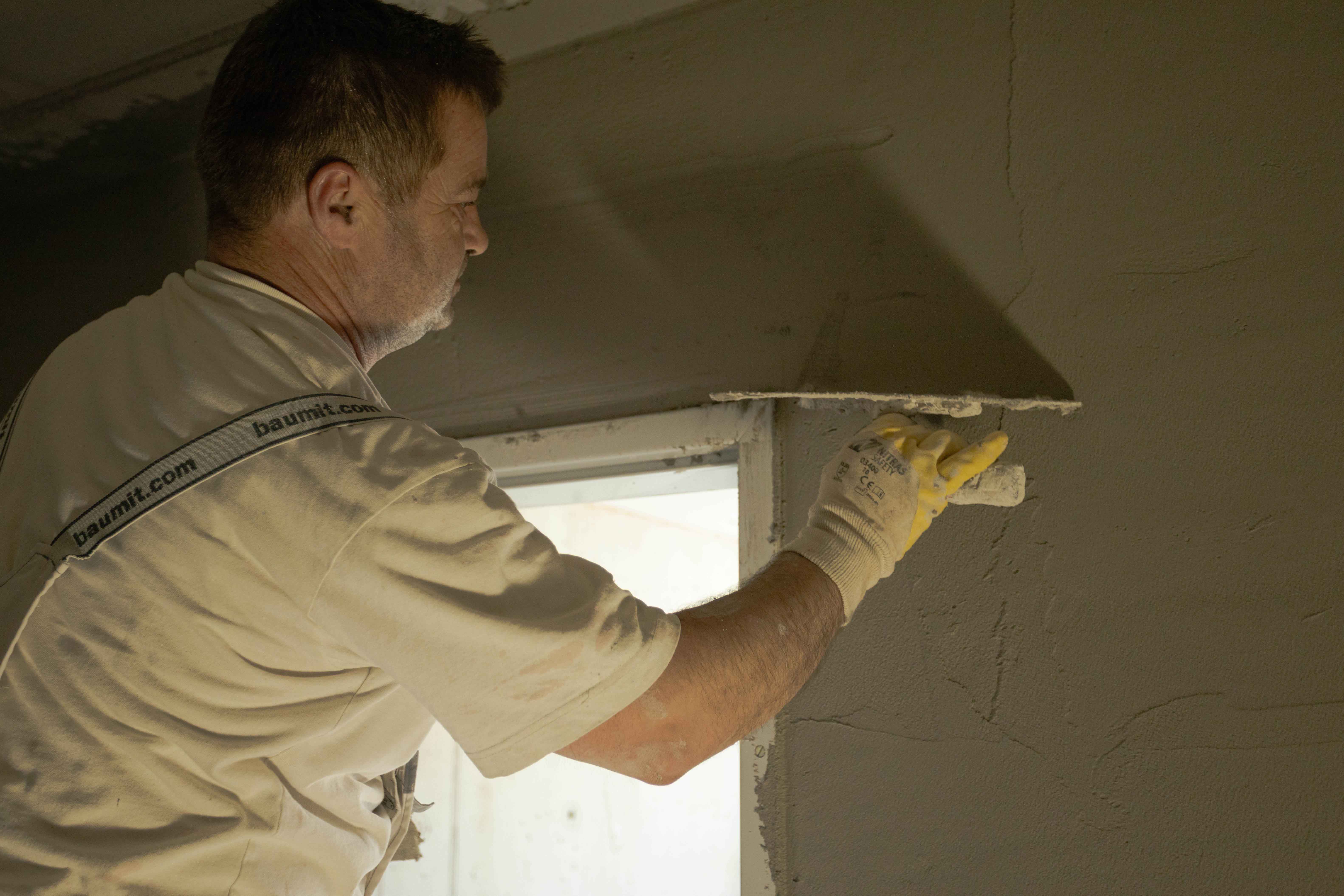 Mann trägt Baustellenkleidung und verputzt eine Wand mit Kelle in einem Raum mit Fenster.