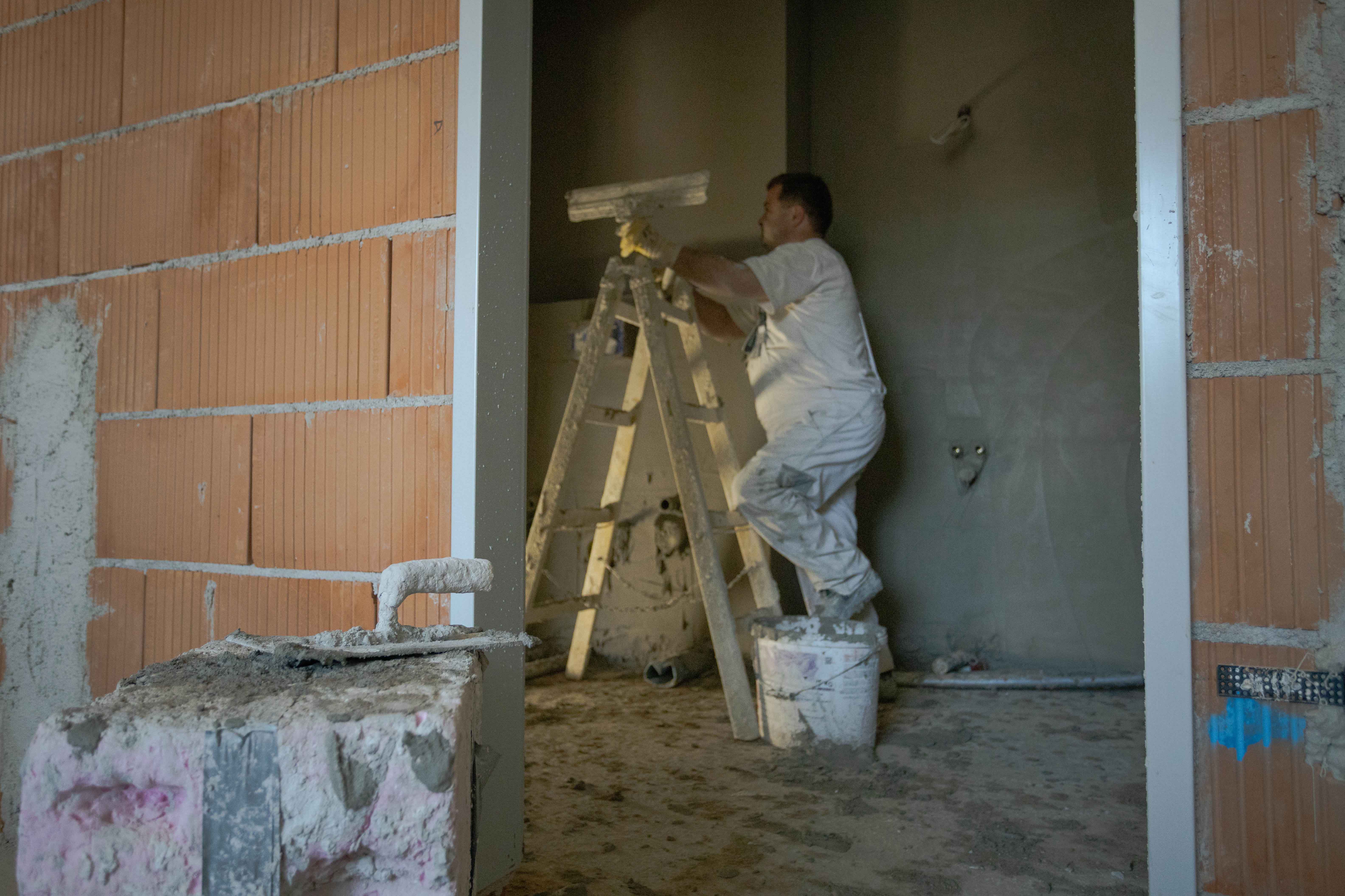 Handwerker steht auf einem Eimer und klettert auf eine Leiter in einem Rohbau mit unverputzten Ziegelwänden.