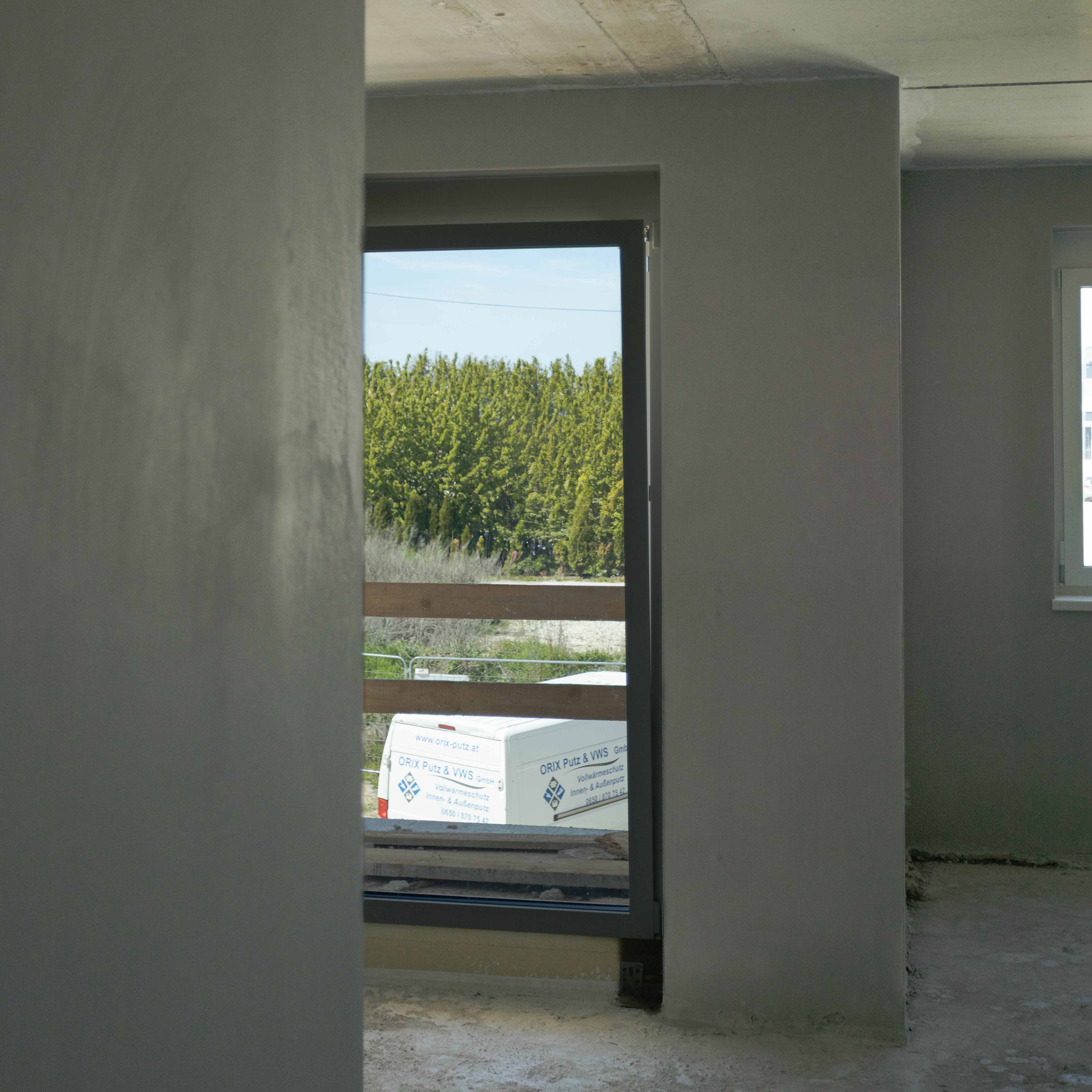 Teil eines unfertigen Innenraums mit Rohbetonboden und Fenster, durch das ein weißer Transporter und grüne Bäume draußen sichtbar sind.