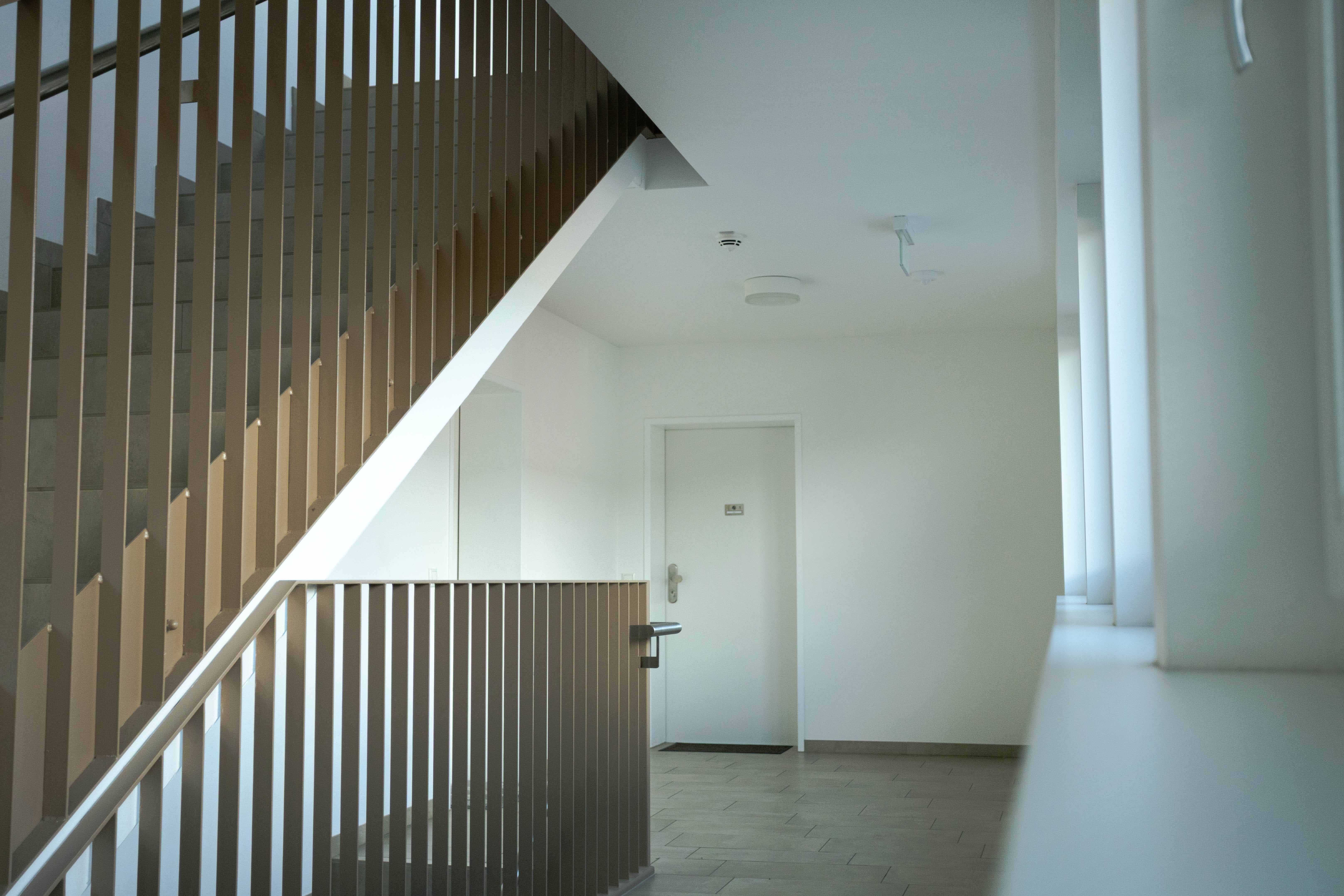 Treppenhaus mit beigen Geländern, weißen Wänden, einer geschlossenen weißen Tür und Fenstern rechts.