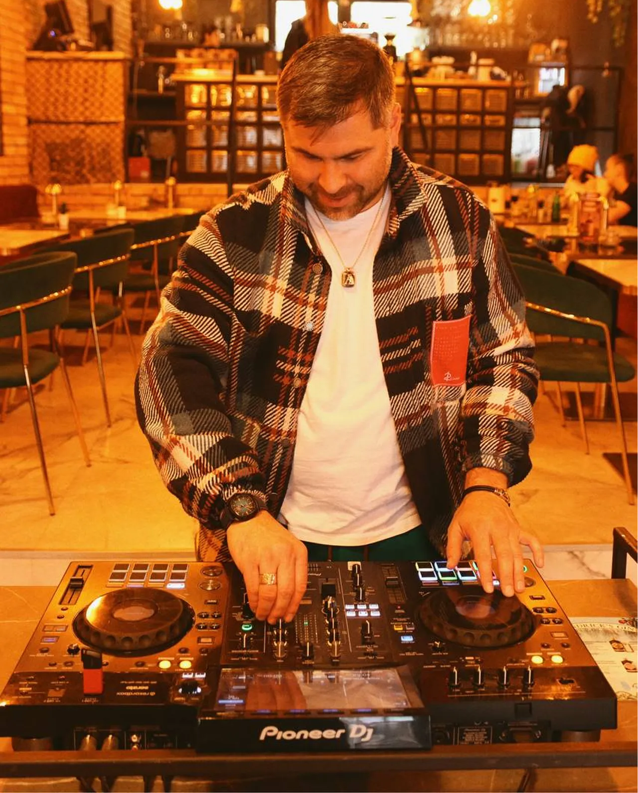 Man wearing plaid jacket and white shirt DJing with a Pioneer DJ controller in a warmly lit cafe.