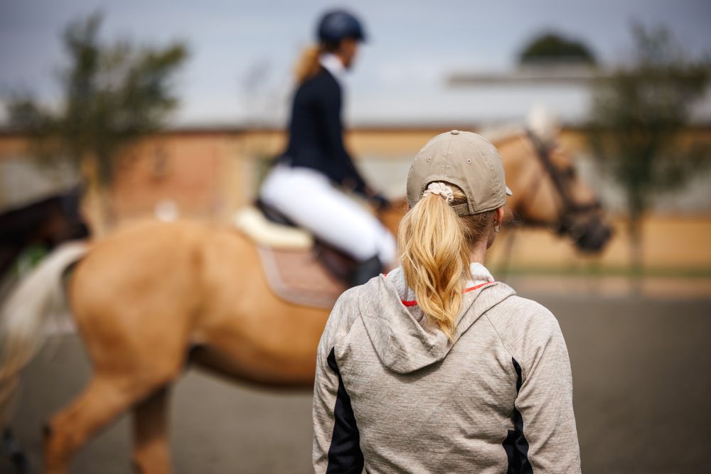 Woman watches a person riding a tan horse in a blurred background.