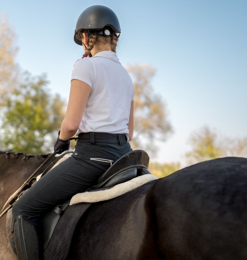Young female equestrian in helmet riding a dark horse outdoors with trees and clear sky behind.