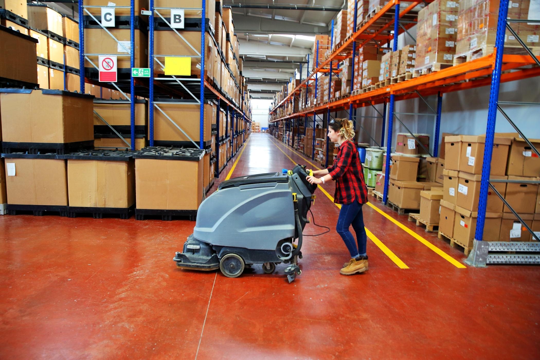 Agent de nettoyage professionnel équipé d'une autolaveuse industrielle dans un atelier de production, sol propre et brillant, éclairage au plafond, équipements de production visibles en arrière-plan.