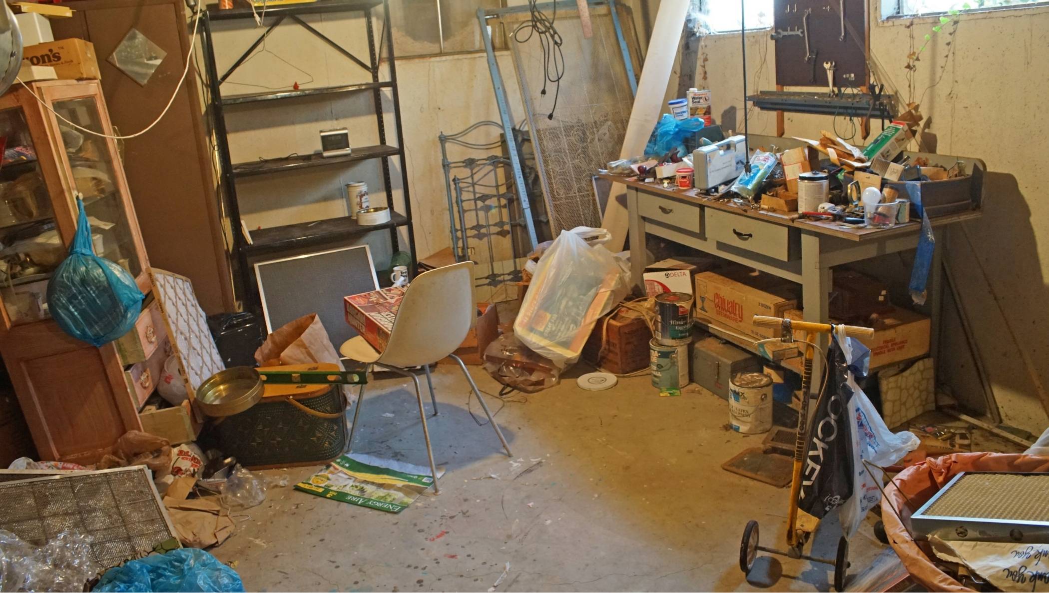 Cave de copropriété encombrée avec cartons, meubles anciens et objets divers empilés, lumière tamisée, ambiance avant-intervention
