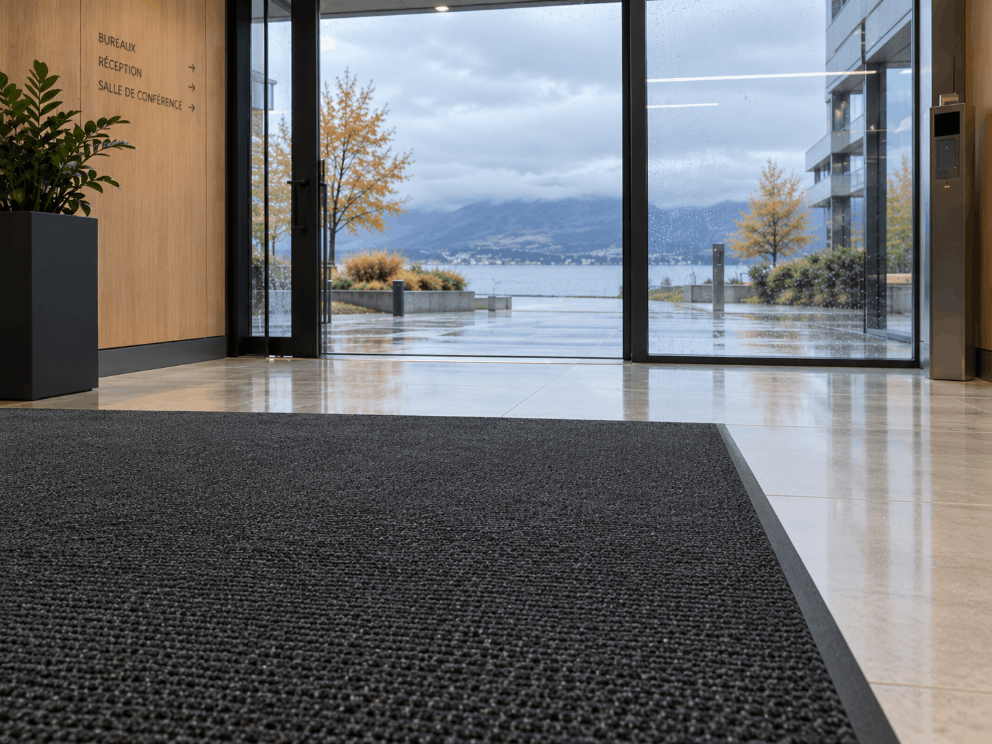 Hall d'entrée d'un immeuble de bureaux contemporain sur la Riviera vaudoise par temps de pluie : grand tapis d'entrée anthracite parfaitement posé, sol clair et sec au-delà du tapis, porte vitrée laissant deviner le lac et un ciel gris d'automne. Lumière naturelle douce, ambiance professionnelle et soignée, cadrage au niveau du sol.