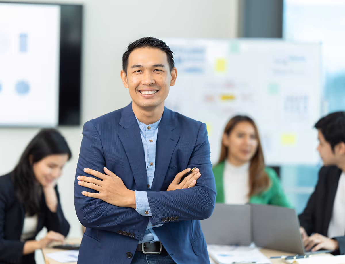 Smiling businessman in a blue suit with arms crossed, with colleagues working on laptops in the background.