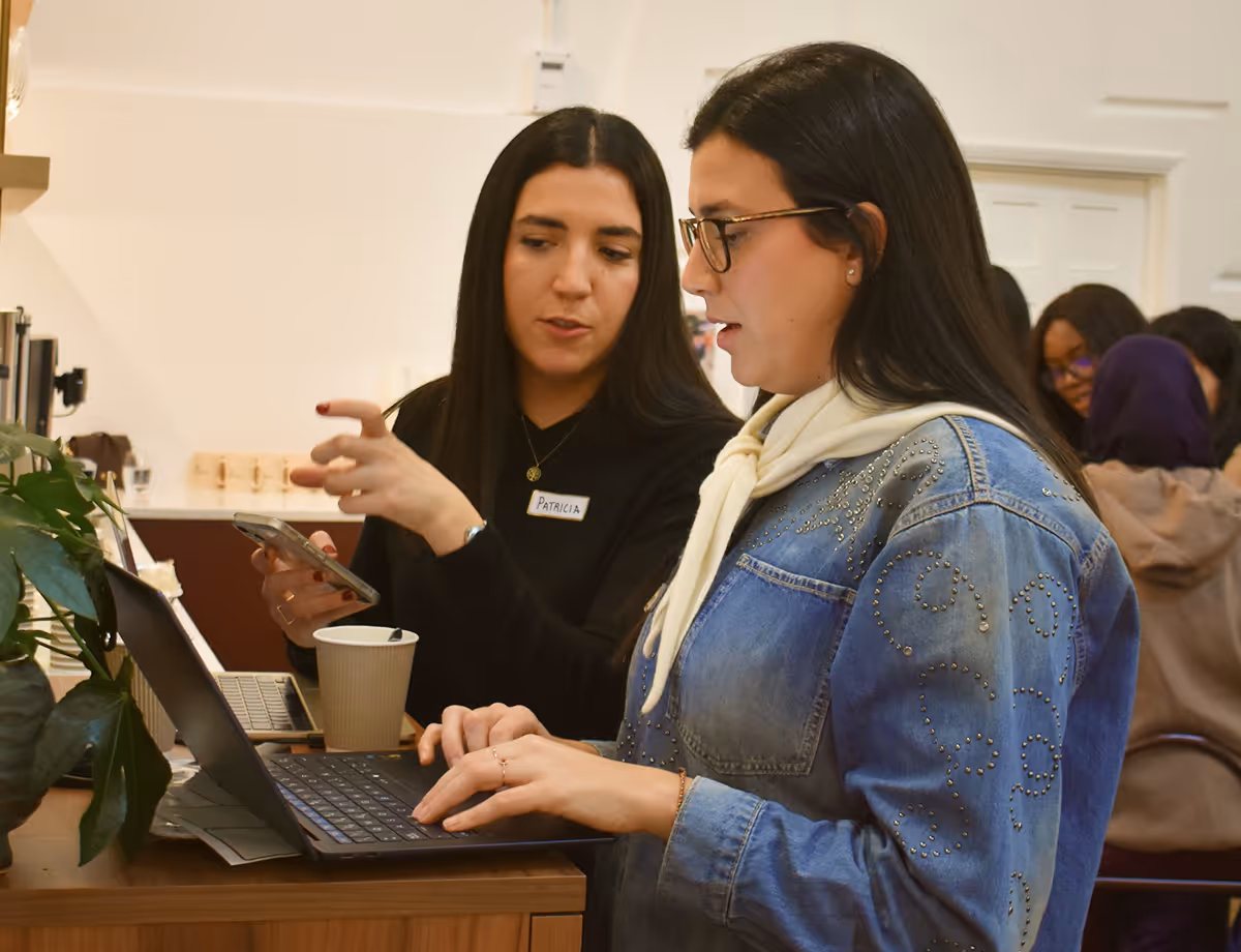 Two women working together at a laptop, one explaining while holding a smartphone and the other typing.