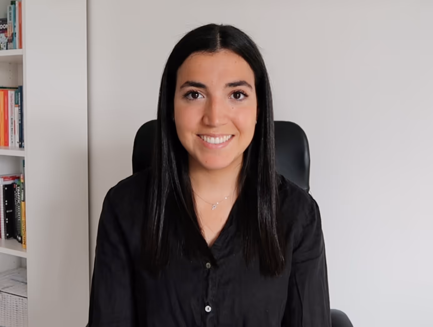 Smiling woman with straight black hair wearing a black shirt, sitting in front of a white wall and bookshelf.