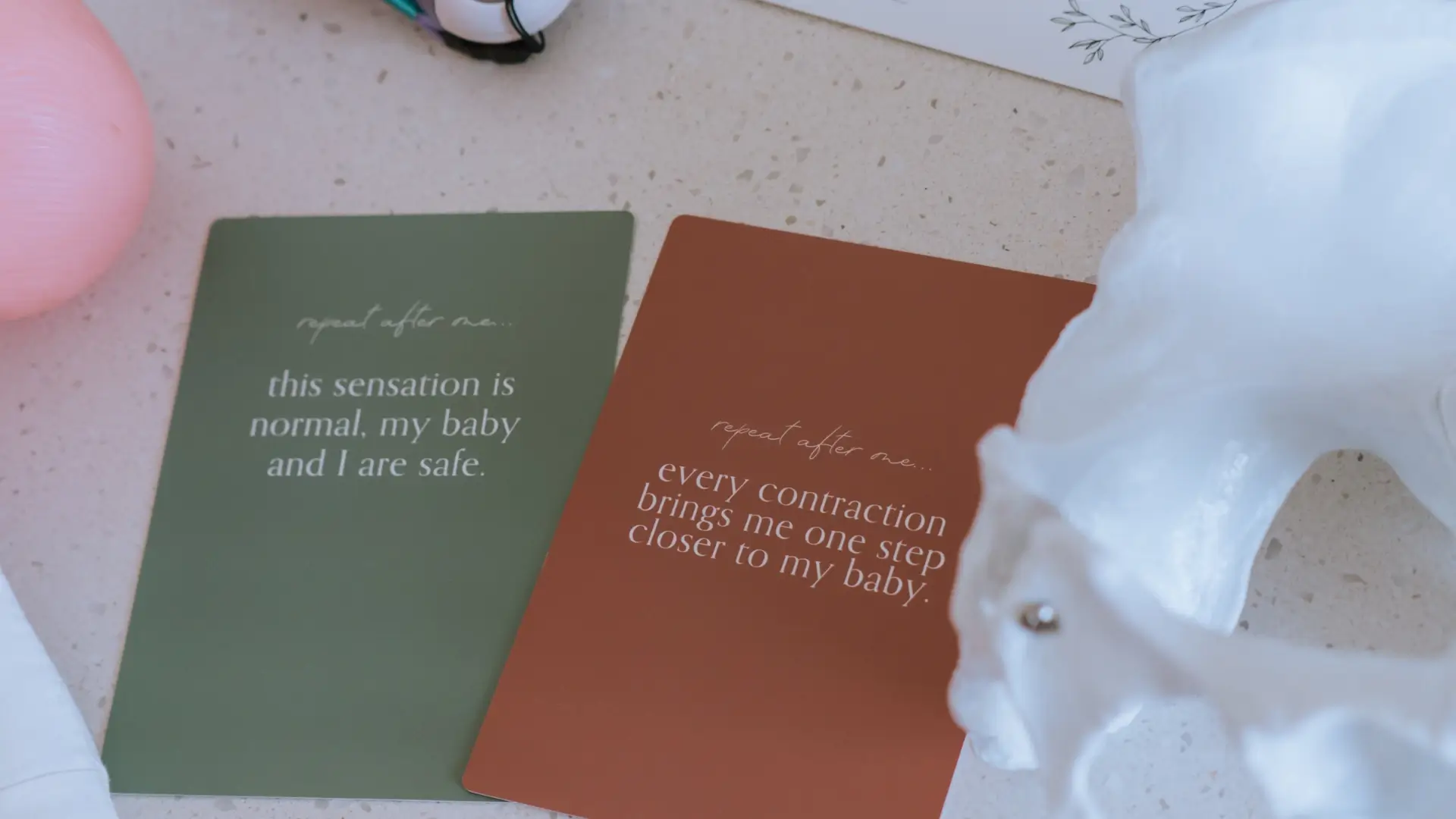 Two cards on a speckled surface with affirmations: one green card says 'this sensation is normal, my baby and I are safe' and one brown card says 'every contraction brings me one step closer to my baby.'
