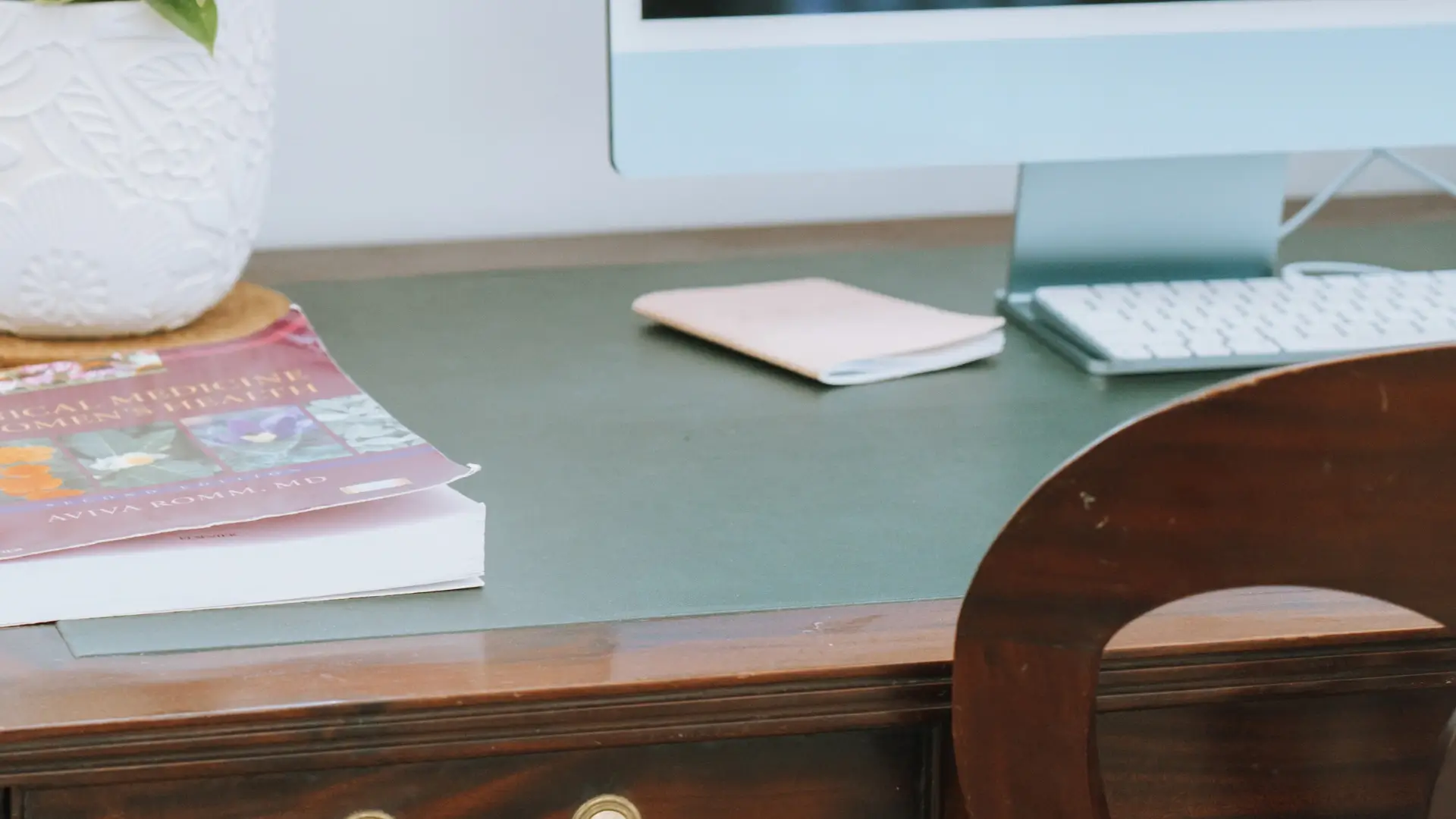 Close-up shot of a wooden office table with an iMac, a vase, a book, and a small notebook on it