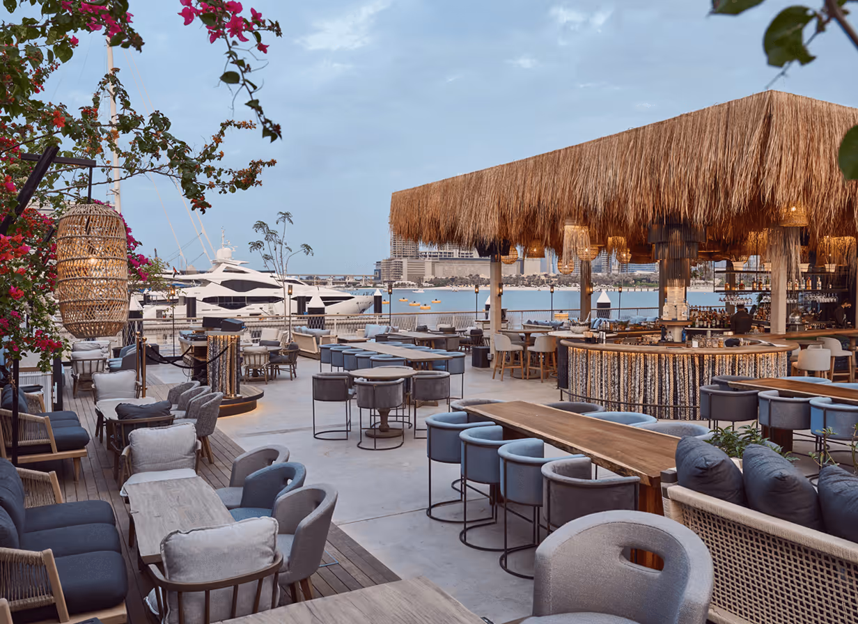 Outdoor waterfront restaurant seating area with modern tables, chairs, a thatched roof bar, and yachts docked in the background.