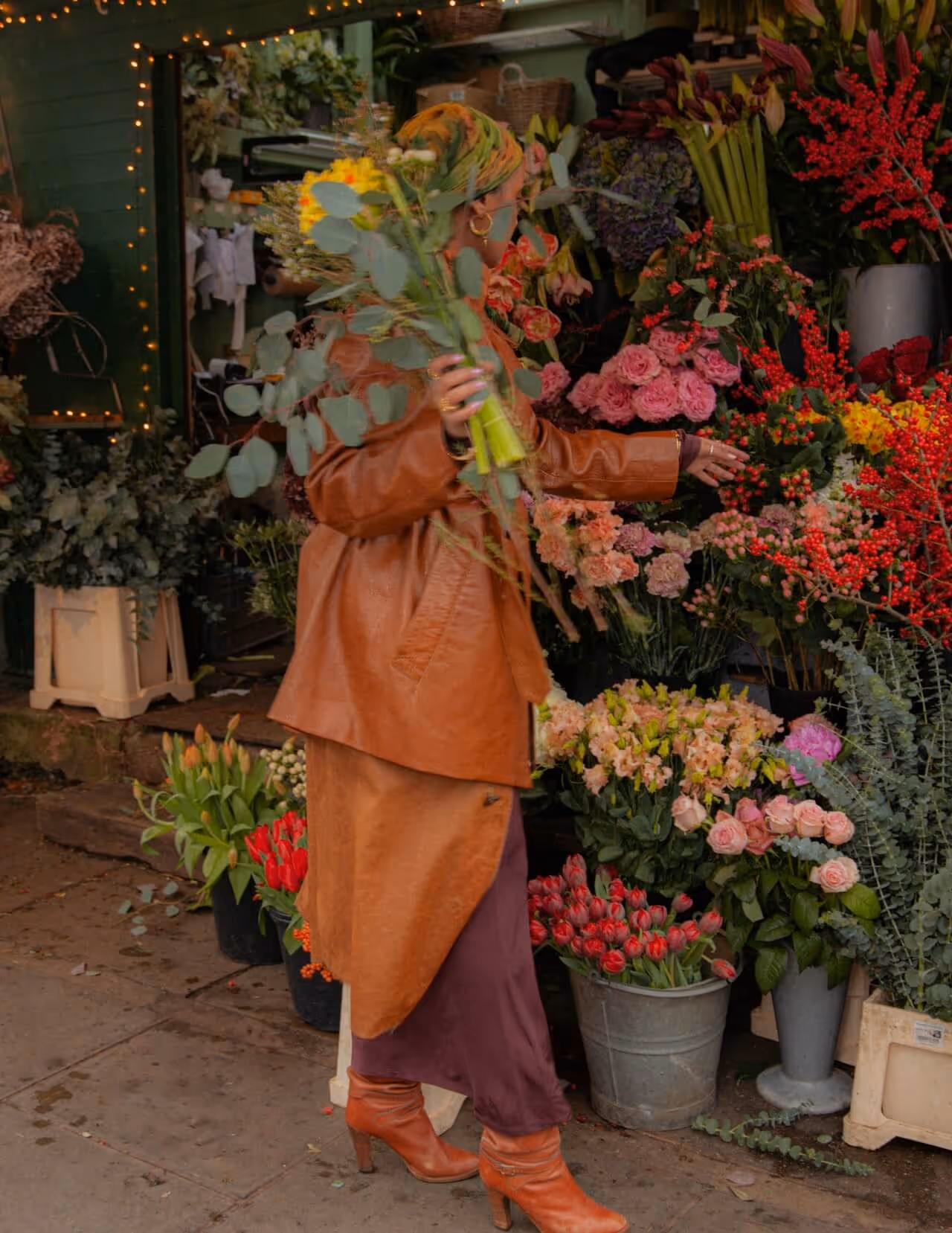 Person wearing a brown leather jacket, purple skirt, and orange boots selecting flowers at an outdoor flower market.