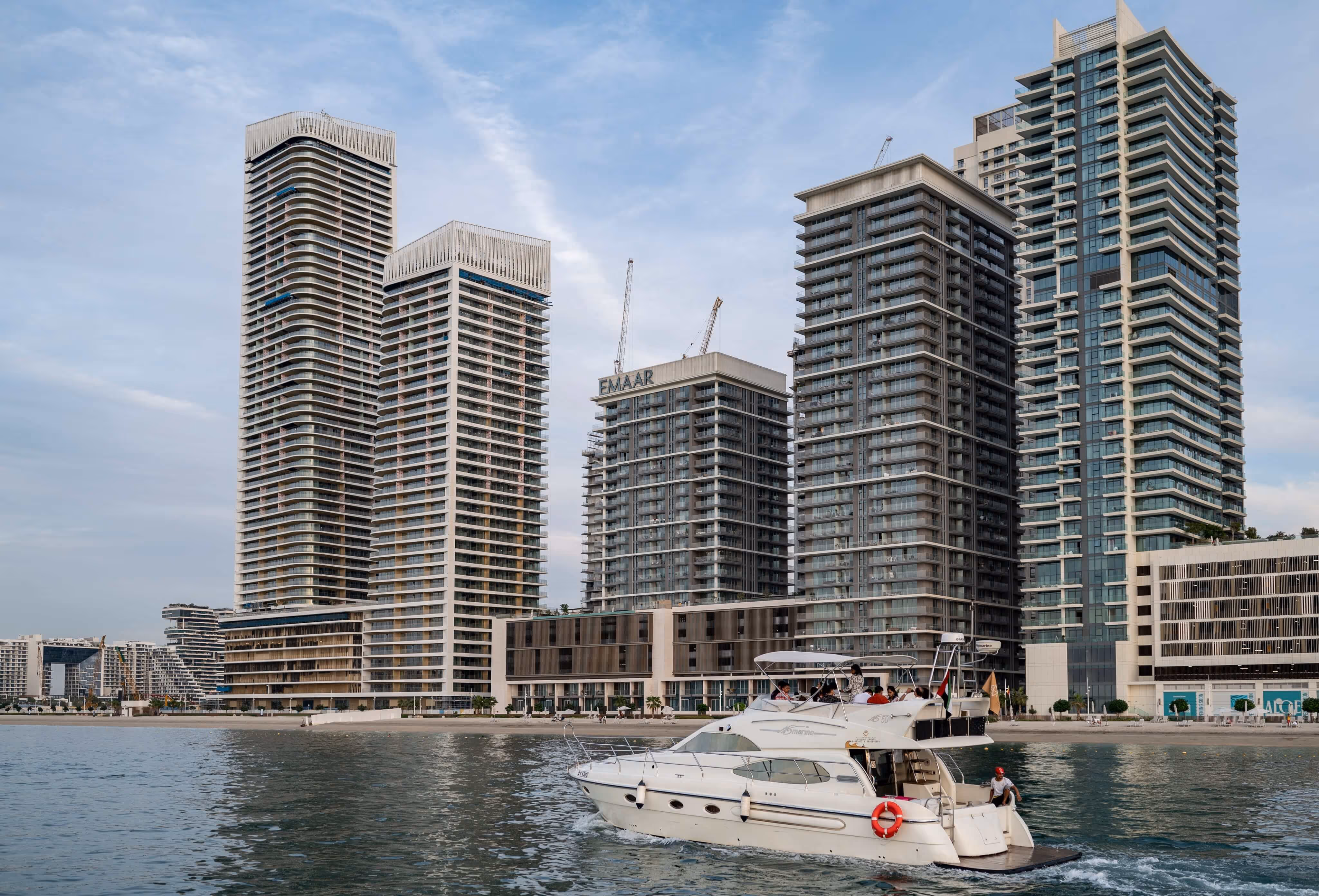 White yacht sailing on calm water with modern high-rise buildings and a sandy shoreline in the background under a partly cloudy sky.