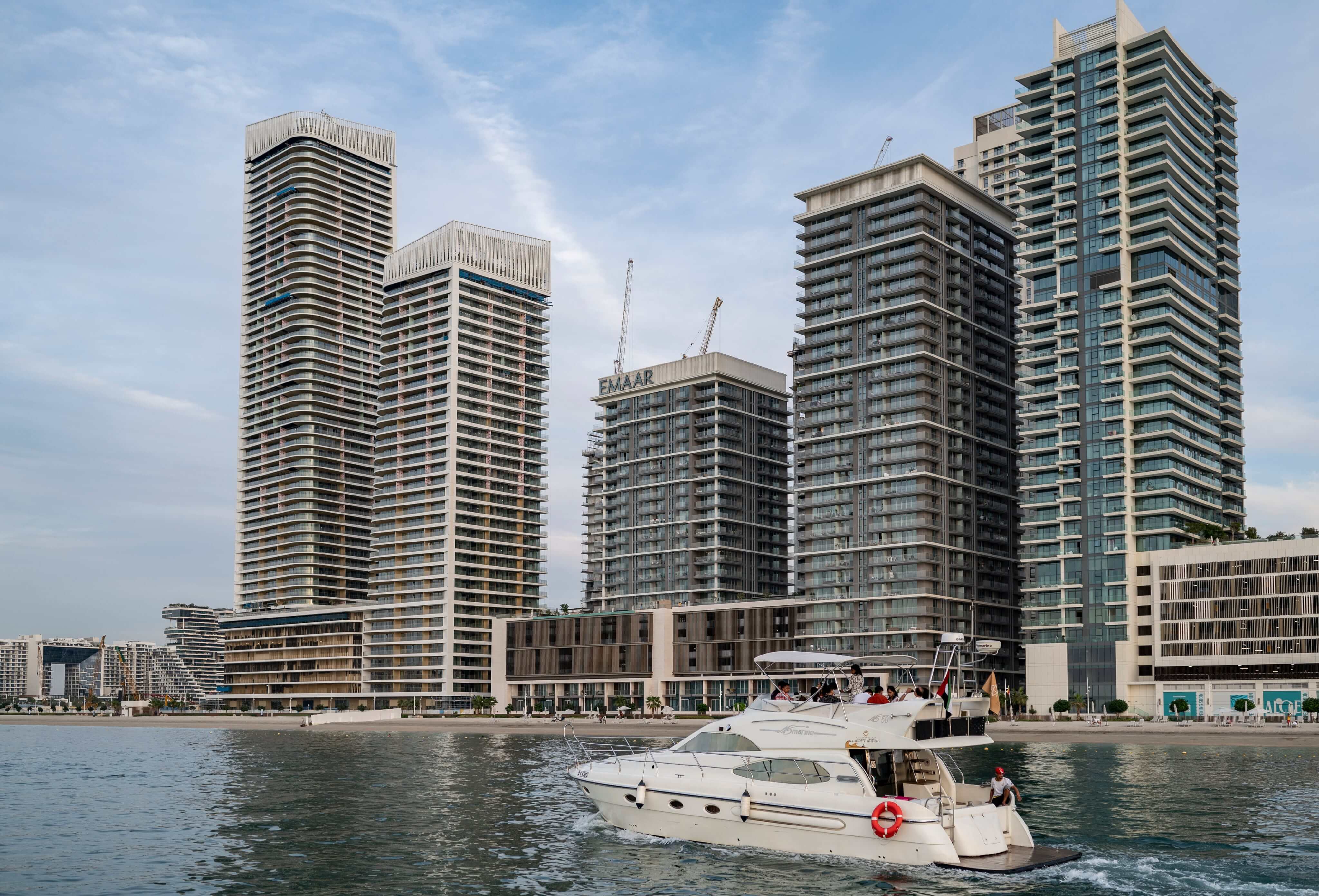White yacht sailing on calm water with modern high-rise buildings and a sandy shoreline in the background under a partly cloudy sky.