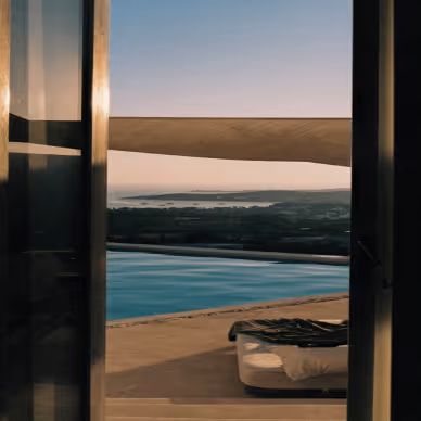 View through open glass doors showing an outdoor pool and a lounge chair with a blanket, overlooking a distant landscape at sunset.