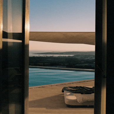 View through open glass doors showing an outdoor pool and a lounge chair with a blanket, overlooking a distant landscape at sunset.