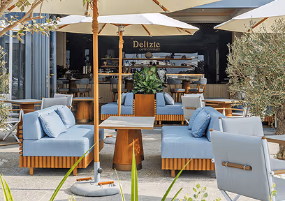 Outdoor seating area with light blue cushioned sofas, wooden tables, and large white umbrellas in front of a café named Delizie.