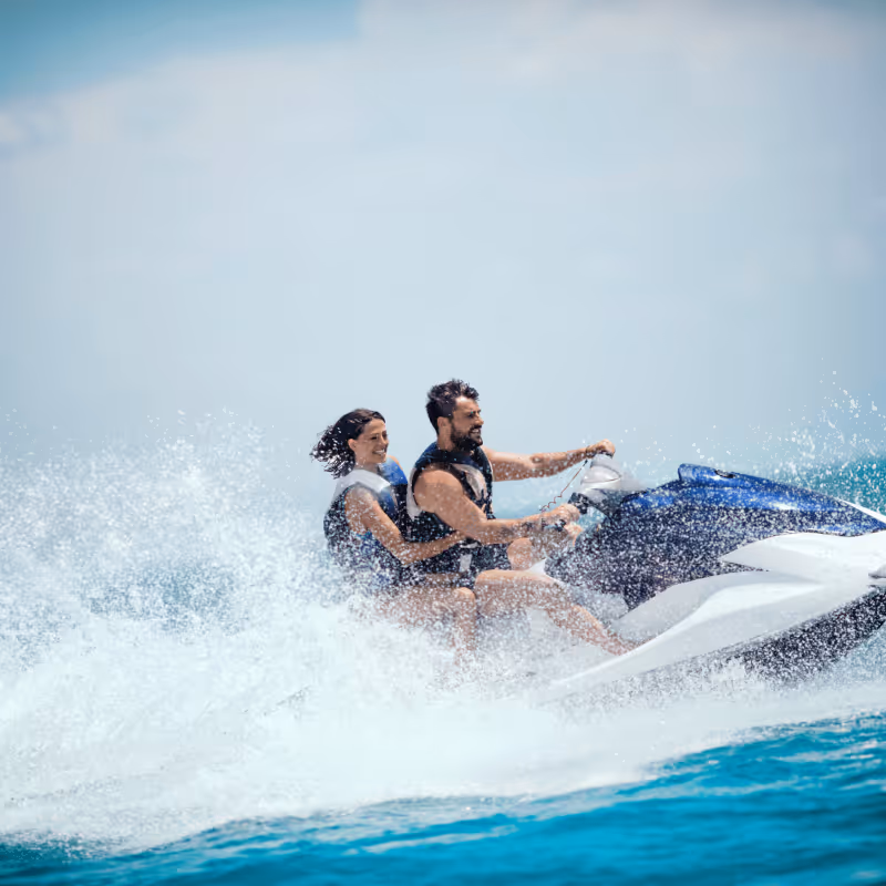 A man and woman wearing life jackets riding a jet ski on blue ocean water with splashing waves.