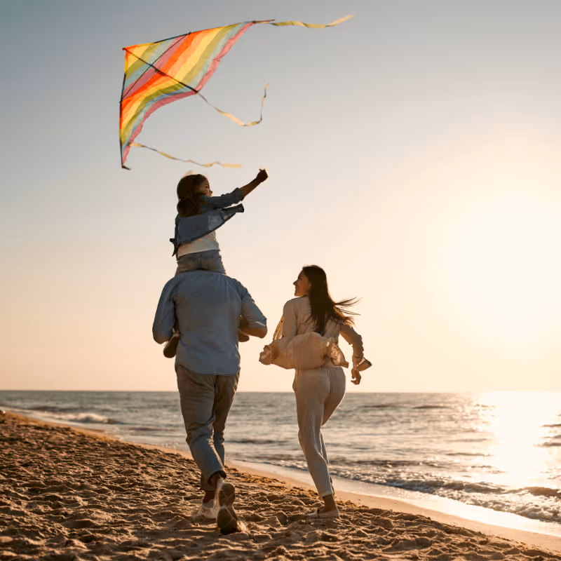 Family running along the beach at sunset with a child flying a colorful kite.