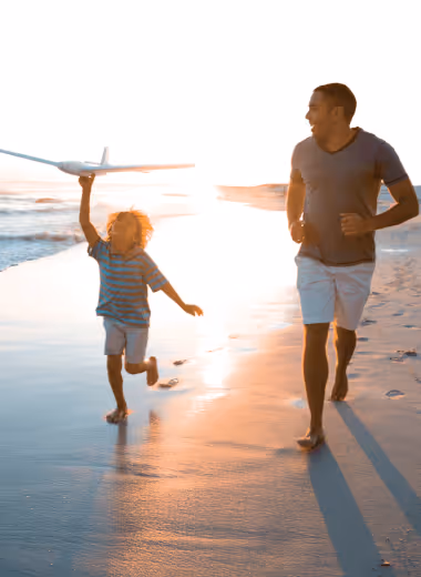 A man and child running barefoot on a sunlit beach, with the child holding a toy airplane overhead.