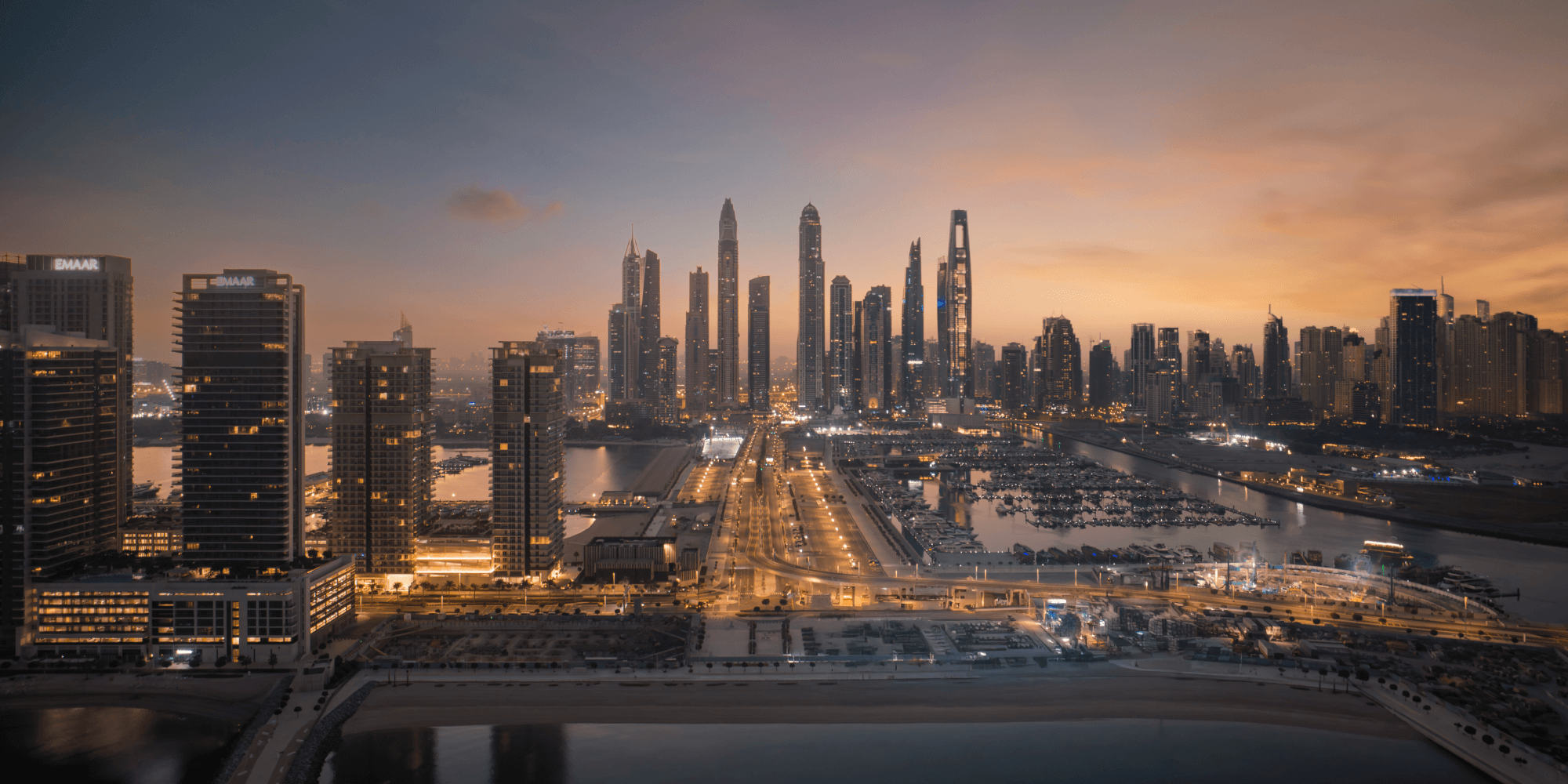 Aerial view of a modern city skyline at dusk with illuminated skyscrapers, roads, and a marina.