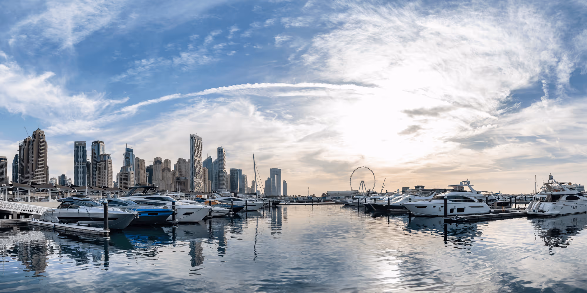 Marina with yachts docked and modern skyscrapers, with a Ferris wheel under a partly cloudy sky.