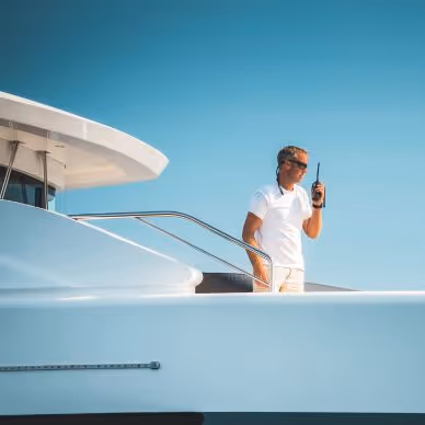 Man in white shirt and sunglasses standing on the deck of a white yacht holding a walkie-talkie under a clear blue sky.
