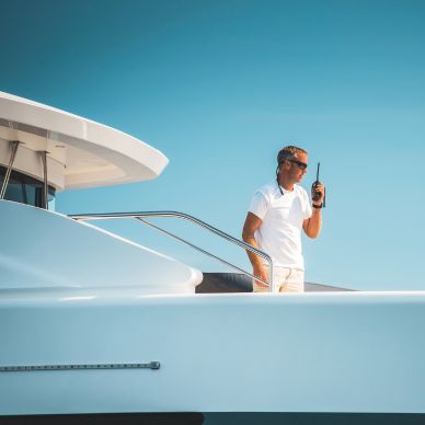 Man in white shirt and sunglasses standing on the deck of a white yacht holding a walkie-talkie under a clear blue sky.
