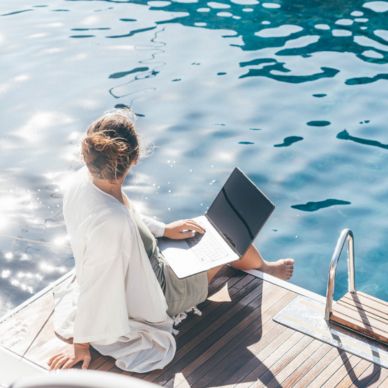 Person sitting on a wooden dock by clear blue water using a laptop and enjoying sunny weather.
