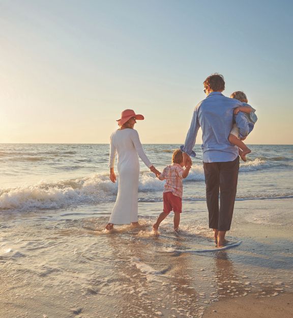 Family of four walking barefoot on the beach near the water during sunset.
