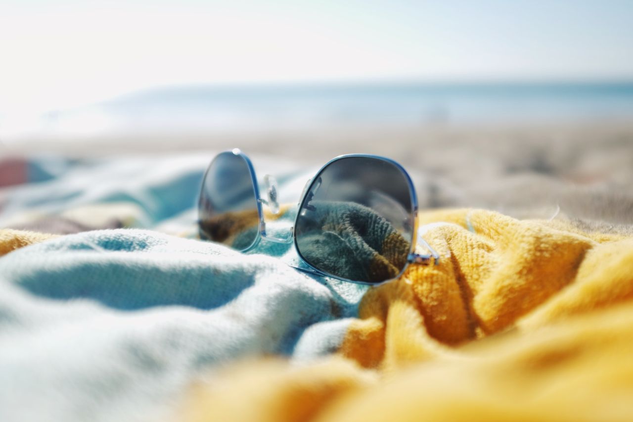Sunglasses resting on a colorful knit blanket with a blurred beach and ocean in the background.