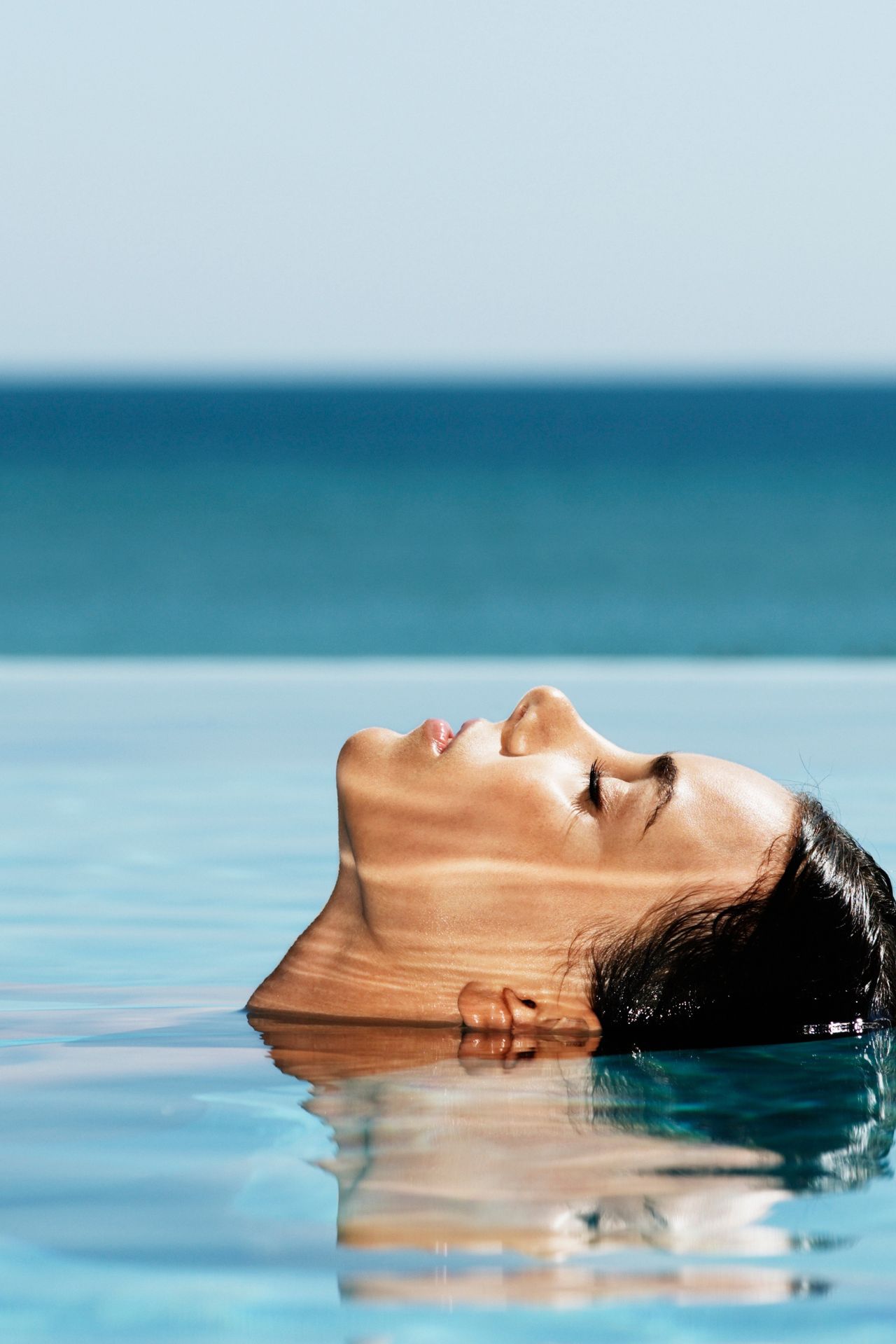 Woman floating peacefully on her back in clear blue water with eyes closed and ocean in the background.