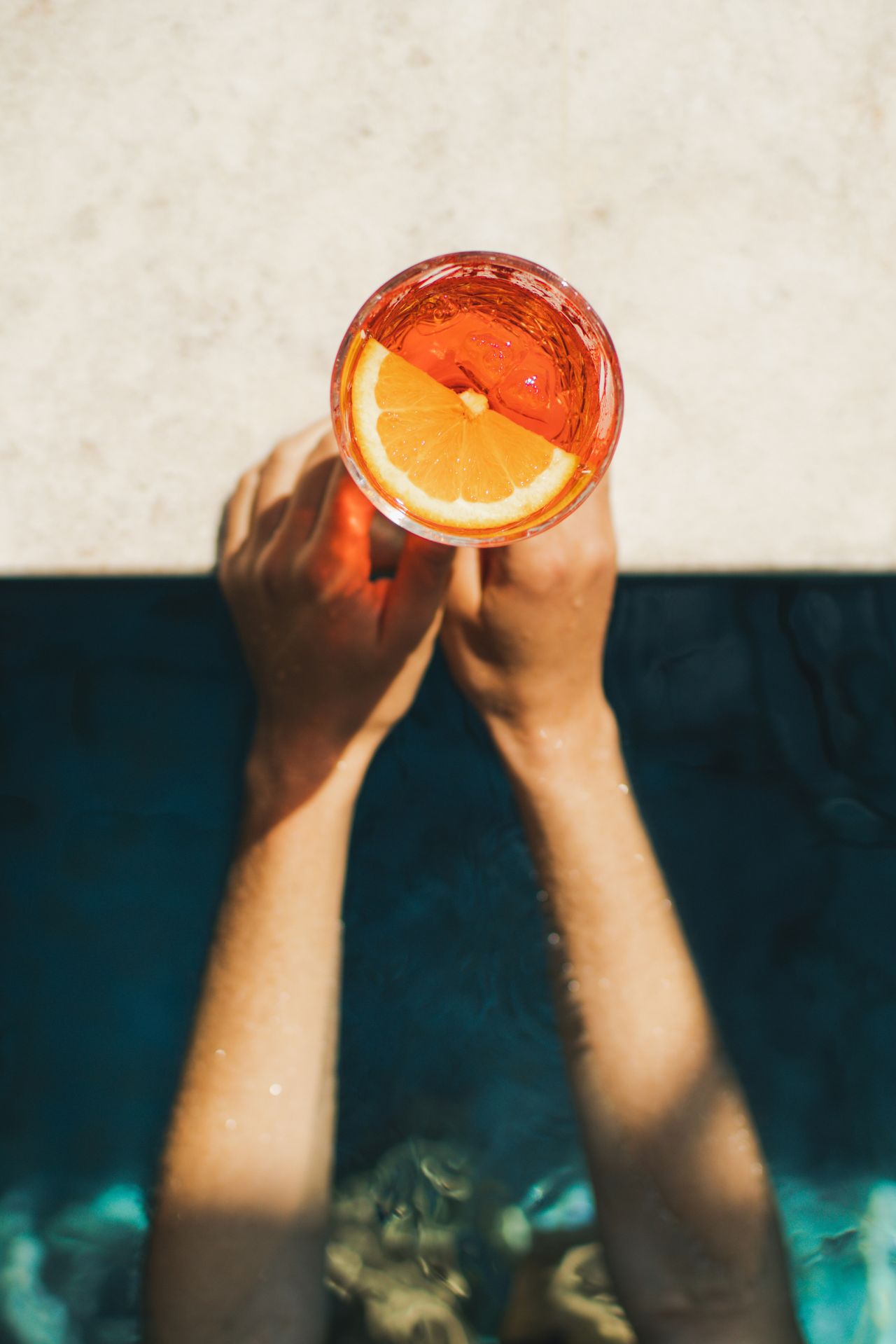 Hands holding a glass with an orange slice and ice over a pool edge.