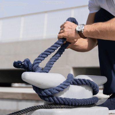 Person tying a thick blue rope around a white dock cleat near water.
